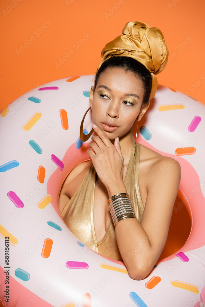 Portrait of pensive african american woman in headscarf posing with pool ring on orange background.