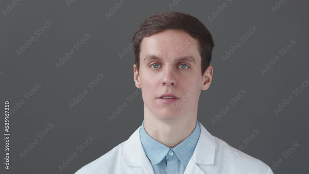 Chest up POV of young male doctor in lab coat speaking at camera on dark grey studio background Chest up POV of young male doctor in lab coat speaking at camera on dark grey studio background