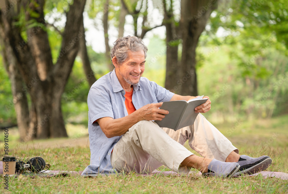 senior man with grey hair holding a book, smiling, sitting near a big tree in forest park. concept of elderly people lifestyle,hobbies,relaxing,health benefits in nature,wellbeing