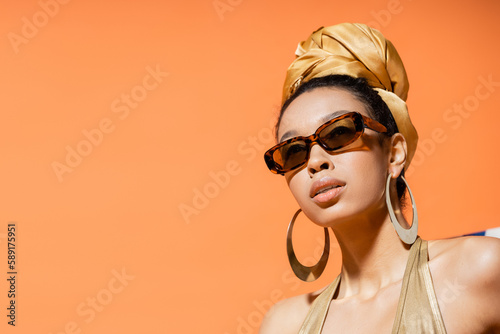 Portrait of fashionable african american model in headscarf and sunglasses isolated on orange.