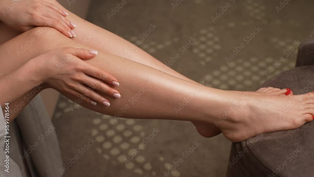Closeup high angle view unrecognizable lady applying moisturizing lotion on leg after hair removal sitting on couch at home. Close up cropped shot of young woman smearing cream on leg at hotel room.
