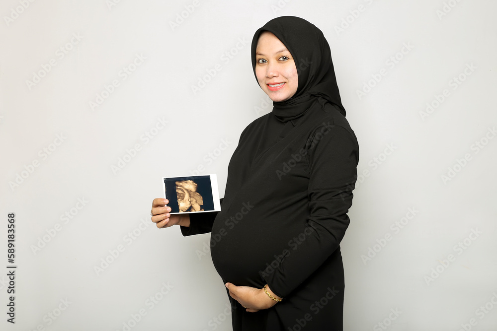 Happy Pregnant Young Hijab Woman holding showing ultrasound scan photo. Pregnant woman hands holding belly on white background.