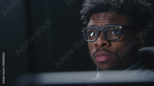 Face close up of African American man working or watching movie in studio with modern design. Film editor looks at monitors. Video footage reflects in the glasses. Freelance and remote work. Zoom in. photo