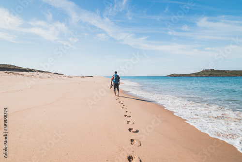 Passionate backpacker, backpack and boots in hand, walks along the Praia da Ilha do Pessegueiro beach on the Atlantic Ocean near Porto Covo, Portugal. In the footsteps of Rota Vicentina