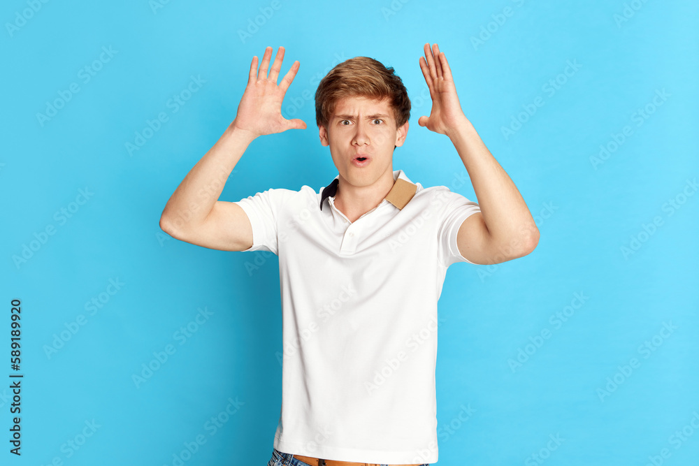 Portrait of young emotive man in casual white t-shirt making hands gestures in annoyance, irritation against blue studio background. Concept of emotions, youth, facial expression, lifestyle