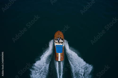 Big luxury wooden motor boat with man and woman moving faster on dark water top view from behind. Large motor boat with people moving on the water, making a white trail, aerial view.