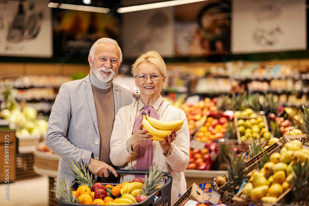 A happy old couple is buying fresh fruits at the supermarket while smiling at the camera.