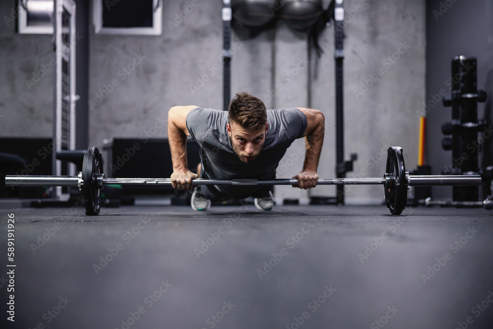 Obraz premium Push-ups and muscle strengthening. An attractive and muscular man does push-ups leaning his hands on a barbell. A man in sportswear pumps his arm muscles in a modern gym. Sport, fitness challenge