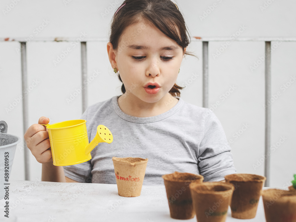 A little caucasian girl holds a yellow watering can with her hands and with surprised emotion looks
