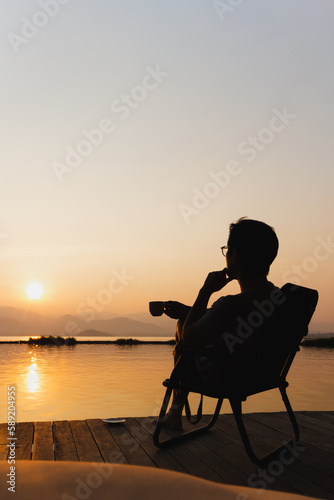 Silhouette tourist man sit on a chair holding cup of coffee wtaching sunrise.