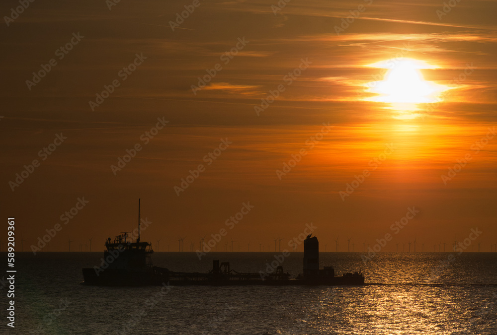 Die Sonne versinkt als glühender Ball wie flüssiges Gold dramatisch in der Nordsee mit Silhouetten von Windkraft Anlagen am Horizont und einem Schiff oder Tanker im Vordergrund