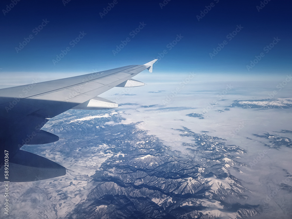 Plane flight above the Carpathian Mountains snowy peaks. Blue skyline and airplane wing seen through the window Plane flight above the Carpathian Mountains snowy peaks. Blue skyline and airplane wing seen through the window