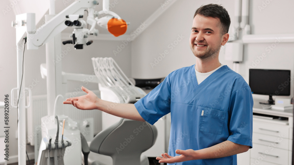 Handsome young dentist in white coat is looking at camera and showing his office Handsome young dentist in white coat is looking at camera and showing his office