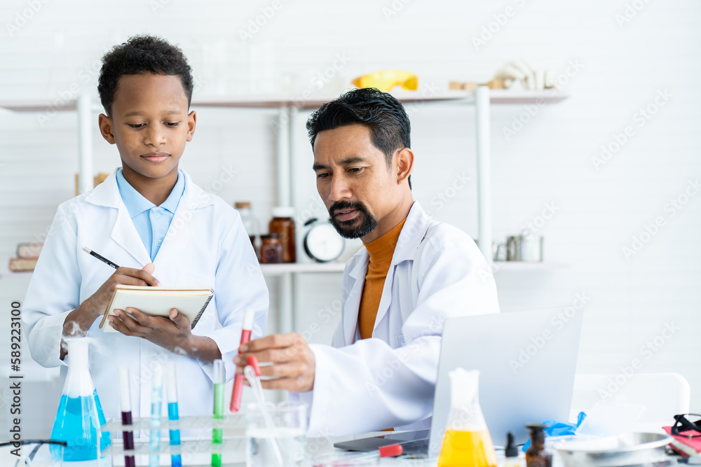 In chemistry classroom with many laboratory tools on table. A young African boy and male teacher in white lab coat doing experiment together. A teacher checking at red test tube and boy writing data. In chemistry classroom with many laboratory tools on table. A young African boy and male teacher in white lab coat doing experiment together. A teacher checking at red test tube and boy writing data.