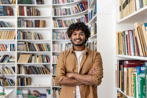 Portrait of happy Hindu student, man with curly hair smiling and looking at camera with arms crossed, student in university academic library.