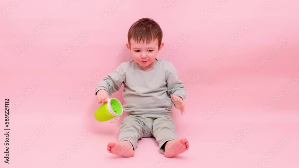Happy toddler baby drinks water from a non-watering glass on a studio pink background. A smiling child with a green no spill plastic cup in his hands. Kid age one year eight months, full height Happy toddler baby drinks water from a non-watering glass on a studio pink background. A smiling child with a green no spill plastic cup in his hands. Kid age one year eight months, full height