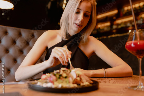 Portrait of young focused woman sitting on brown sofa at wooden table near black plate with vegetable salad and glass with red cocktail, holding fork, eating in restaurant cafe. Food. Soft focus.