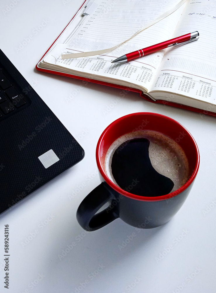 Black-red mug of coffee black laptop red planner and red pen on the white desk Black-red mug of coffee black laptop red planner and red pen on the white desk