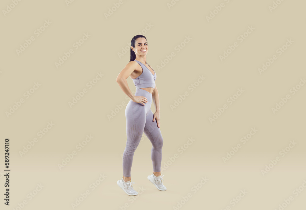 Obraz premium Full-length portrait of smiling active woman athlete in sportswear posing on nude brown studio background. Toned fit girl coach or trainer show good body shape. Fitness and sport concept.