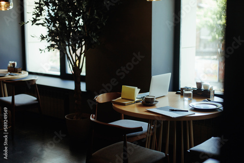 Empty round brown wooden table near window in cafe with open laptop, cup of coffee, open book, plate, black menu with pen. Technology, internet, business, distance work, freelancing. Copy space.