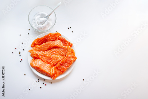 Fresh raw salmon fish fillet on a white plate, a small bowl of salt and black peppercorns on white background. Healthy food. 
