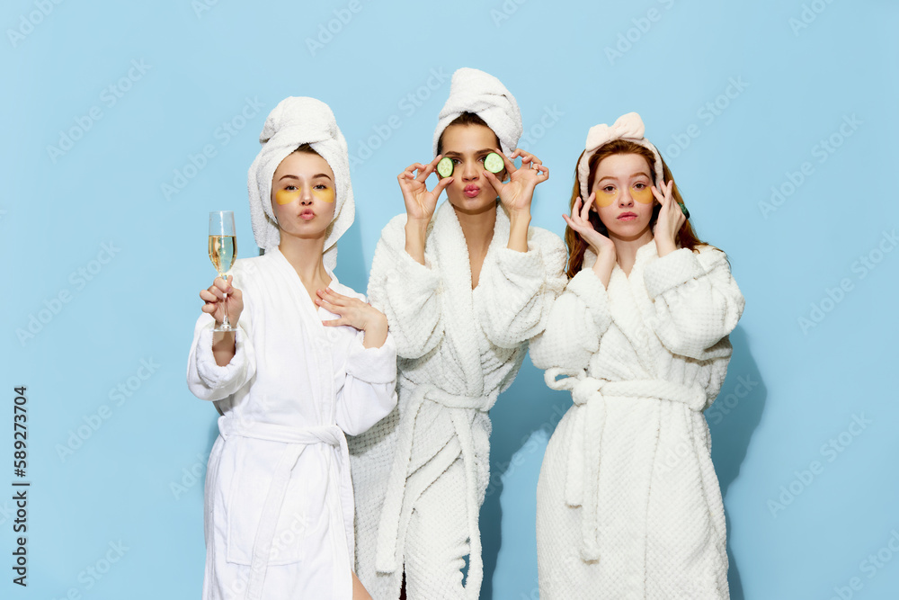 Attractive young girls in bathrobes and face masks taking care after skin, getting readyfor party with champagne against blue background. Concept of youth, face care, beauty, friendship, relaxation - obrazy, fototapety, plakaty 