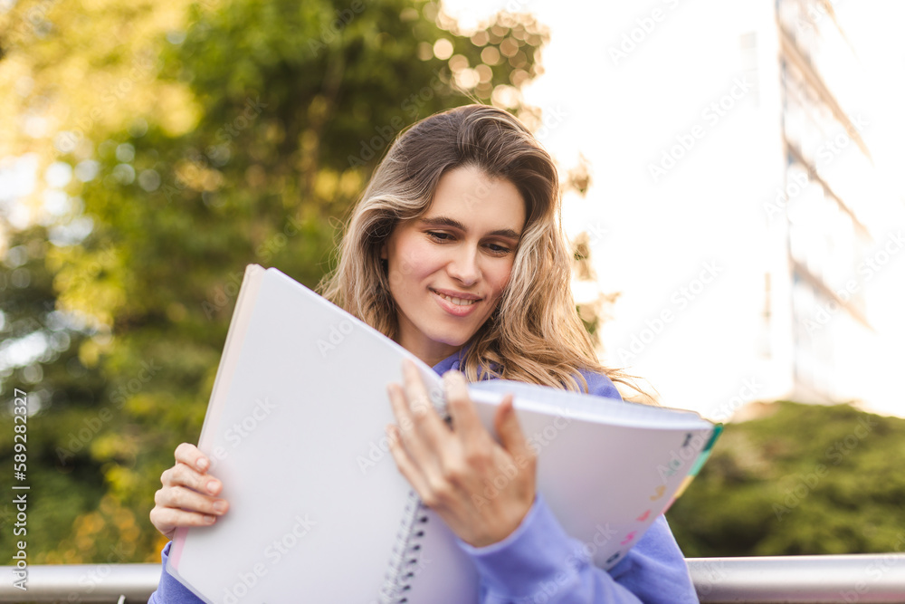 Attractive student woman holding personal diary sitting on bench in summer park, look happy and smiling. Blonde curly girl wear purple hoody, skirt, hold pink notebook and read it, pink bag on bench. Attractive student woman holding personal diary sitting on bench in summer park, look happy and smiling. Blonde curly girl wear purple hoody, skirt, hold pink notebook and read it, pink bag on bench.