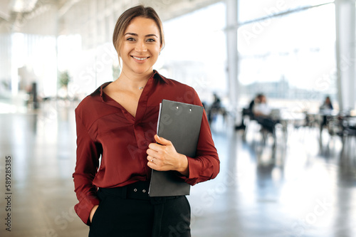 Portrait of a gorgeous successful confident positive blonde caucasian business lady dressed in an elegant wear, stands in a business center, holds documents, looks at camera, smiling friendly photo