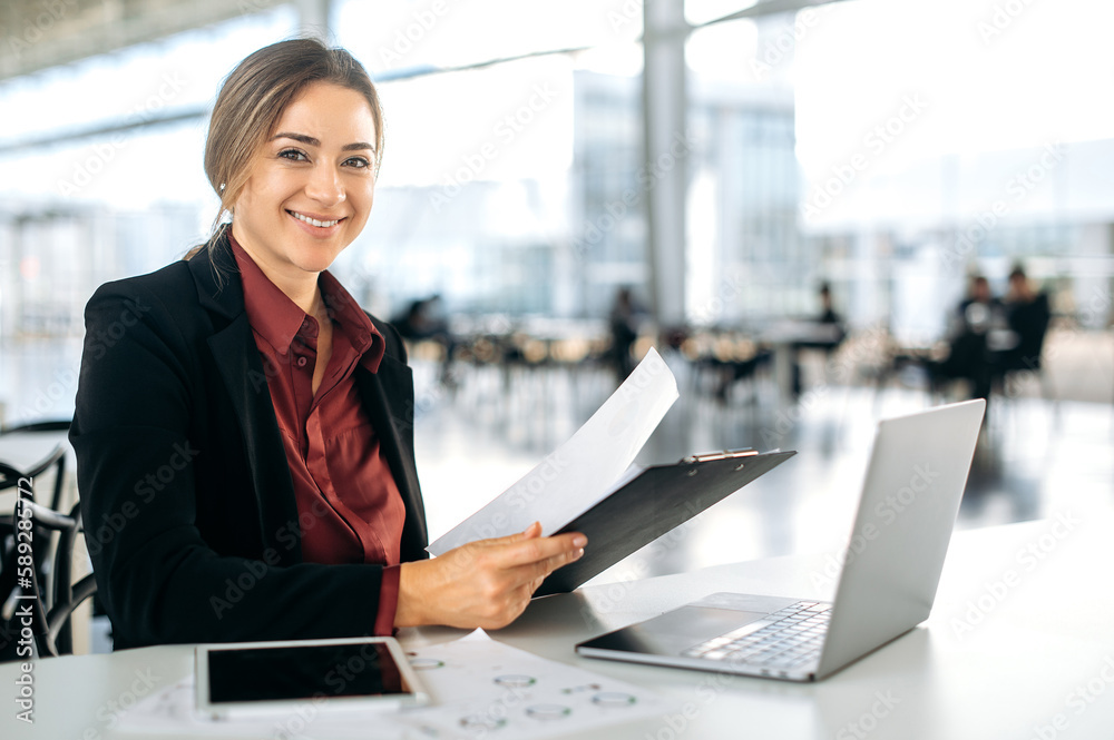 Photo of a beautiful positive successful caucasian business lady sitting at her desk in a modern office, working with documents and a laptop on a project, looking at the camera, smiling friendly Photo of a beautiful positive successful caucasian business lady sitting at her desk in a modern office, working with documents and a laptop on a project, looking at the camera, smiling friendly