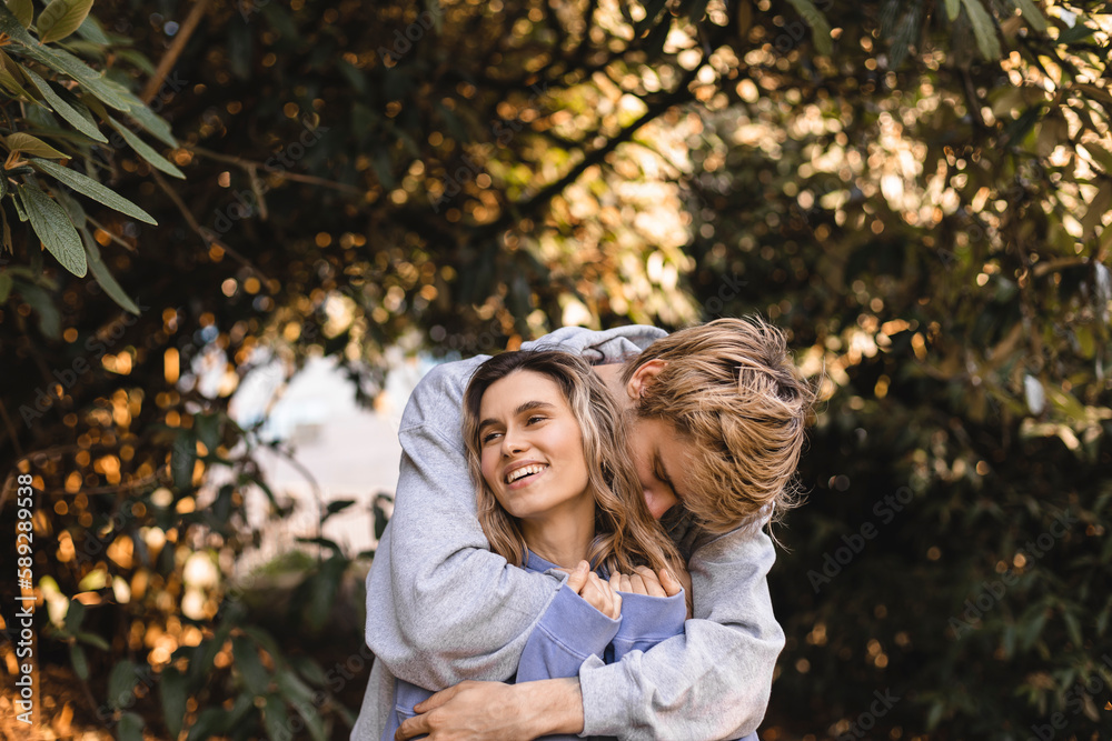 Fototapeta premium Portrait of beautiful blonde curly girl looking away with happy smile while man kissing her neck. Happy couple walk in park.