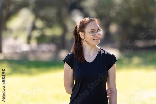 Fototapeta Naklejka Na Ścianę i Meble -  An adult smiling woman wearing glasses and sports clothes standing in the park