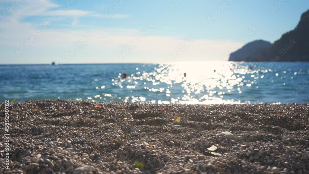 Closeup detail - small pebbles or rough sand on a beach, blurred afternoon sun lit calm sea in background, camera pan slowly, blurred people walking at water distance Closeup detail - small pebbles or rough sand on a beach, blurred afternoon sun lit calm sea in background, camera pan slowly, blurred people walking at water distance