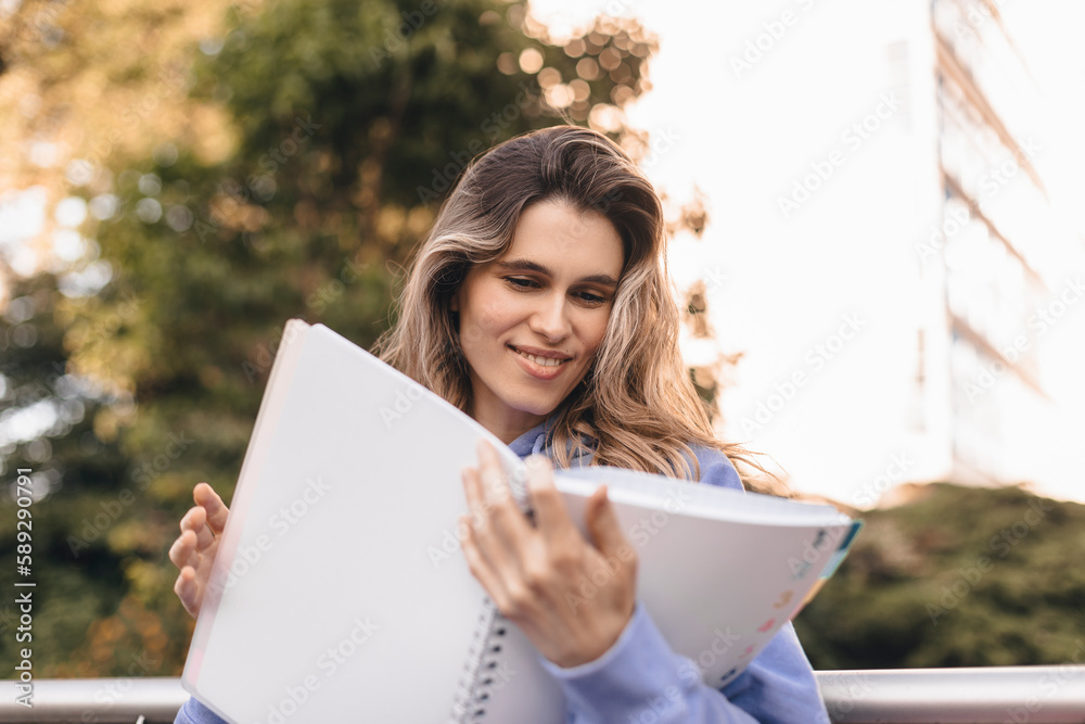 Attractive student woman holding personal diary sitting on bench in summer park, look happy and smiling. Blonde curly girl wear purple hoody, skirt, hold pink notebook and read it, pink bag on bench. Attractive student woman holding personal diary sitting on bench in summer park, look happy and smiling. Blonde curly girl wear purple hoody, skirt, hold pink notebook and read it, pink bag on bench.