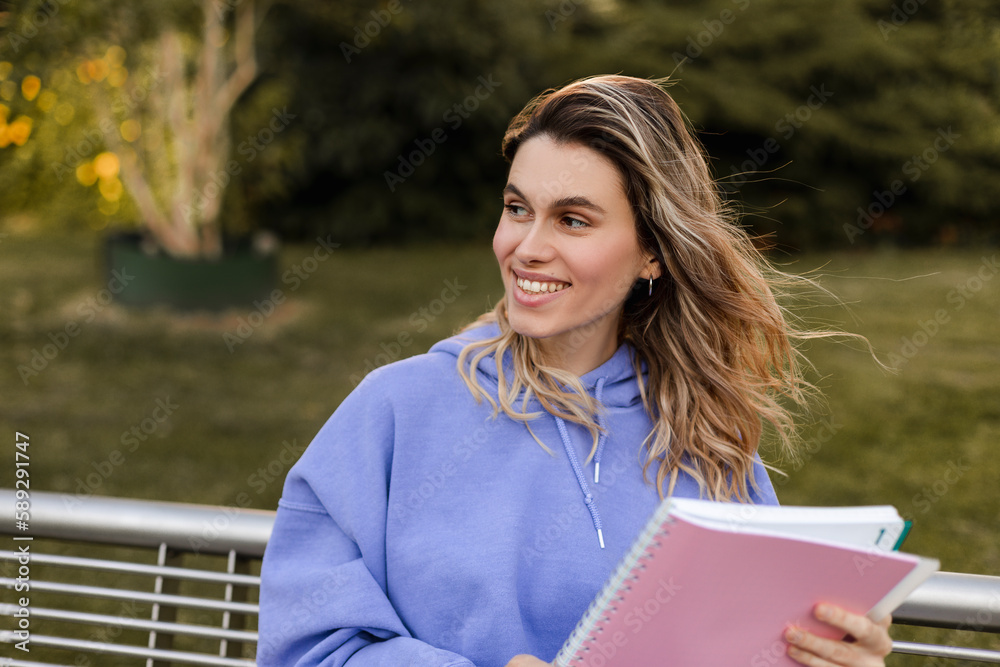 Happy blonde curly woman in purple hoody, hold pink sketchbook, sitting on bench in the park. Girl look at side and look happy, smiling. Rest or studying in the park. Happy blonde curly woman in purple hoody, hold pink sketchbook, sitting on bench in the park. Girl look at side and look happy, smiling. Rest or studying in the park.