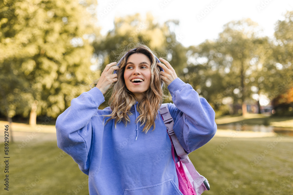 Attractive curly blonde woman walk on the city park street. Girl wear purple hoodie, pink bag and look happy and smiles. Woman put on hood while go on the street, good mood and day.