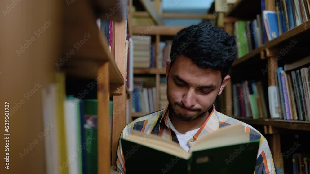 University Library: Talented Indian Boy Standing Next to Bookshelf Reads Book for His Class Assignment and Exam Preparations University Library: Talented Indian Boy Standing Next to Bookshelf Reads Book for His Class Assignment and Exam Preparations