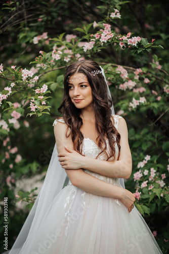 A beautiful bride with long curly hair in a chic dress, smiling, holding her wedding bouquet, looking into the lens. Portrait of the bride. Spring wedding. Natural makeup