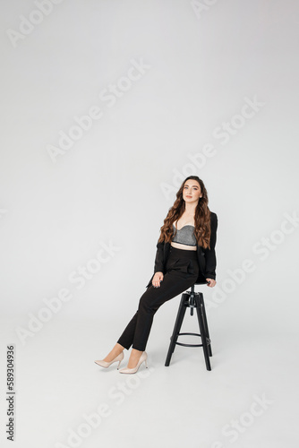 young girl with dark curly hair in a suit sits on a high chair on a white background, copy space © sadreevaa