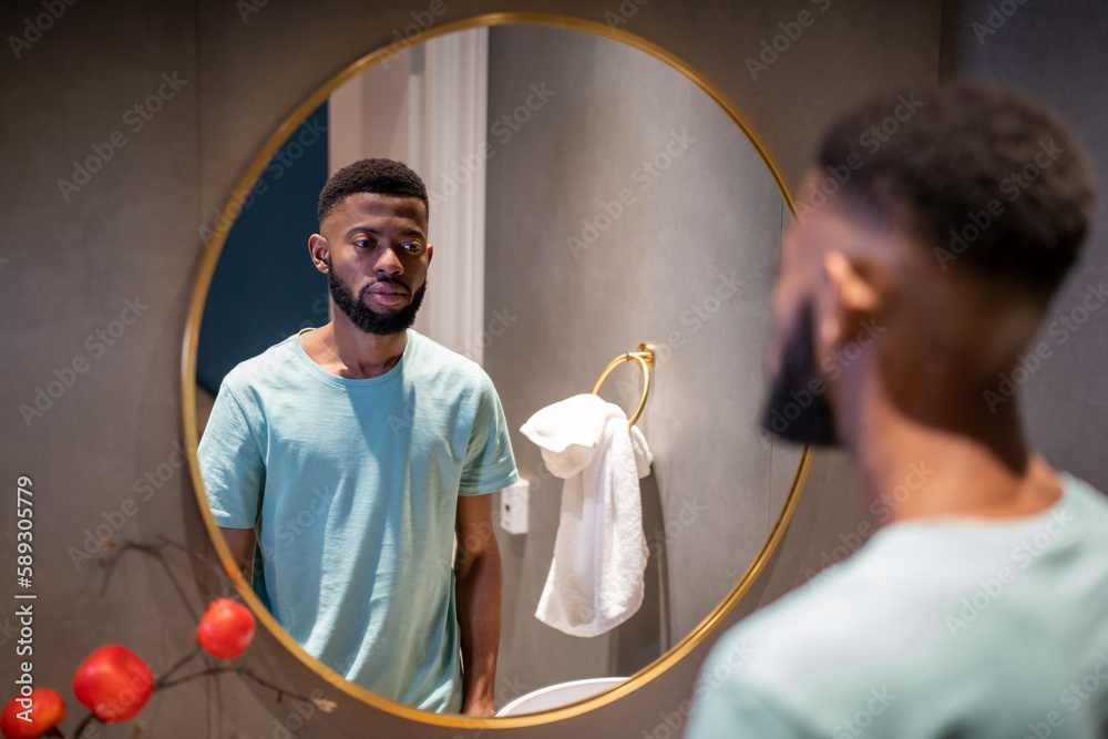 Unhappy upset 30s African American man looking in mirror, standing in