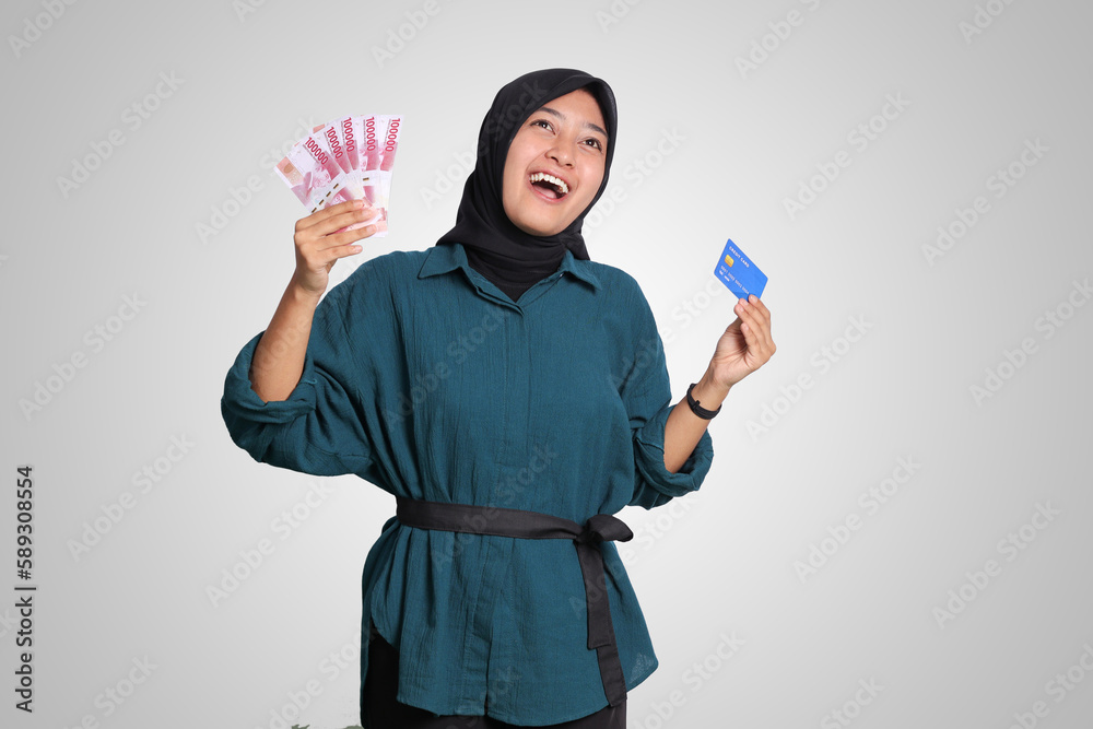 Portrait of cheerful Asian muslim woman with hijab, showing one hundred thousand rupiah while holding a credit card. Financial and savings concept. Isolated image on white background Portrait of cheerful Asian muslim woman with hijab, showing one hundred thousand rupiah while holding a credit card. Financial and savings concept. Isolated image on white background