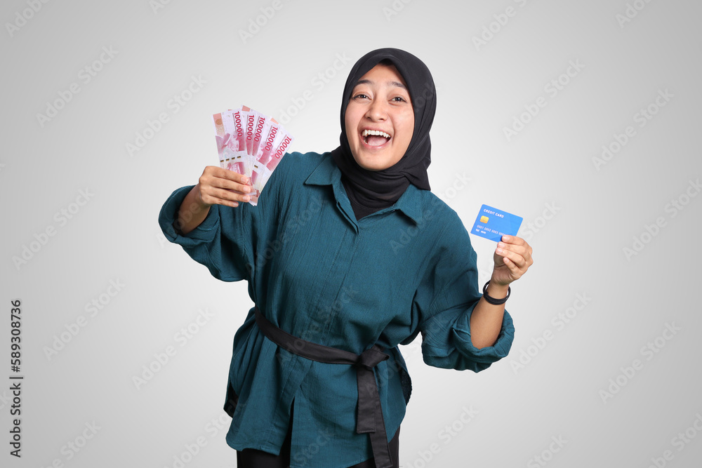 Portrait of cheerful Asian muslim woman with hijab, showing one hundred thousand rupiah while holding a credit card. Financial and savings concept. Isolated image on white background Portrait of cheerful Asian muslim woman with hijab, showing one hundred thousand rupiah while holding a credit card. Financial and savings concept. Isolated image on white background