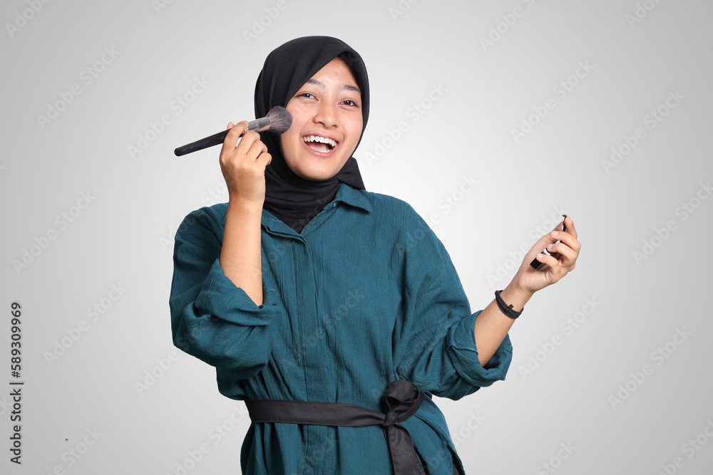 Portrait of excited Asian muslim woman with hijab, applying foundation powder or blush with makeup brush on her cheek. Isolated image on white background Portrait of excited Asian muslim woman with hijab, applying foundation powder or blush with makeup brush on her cheek. Isolated image on white background
