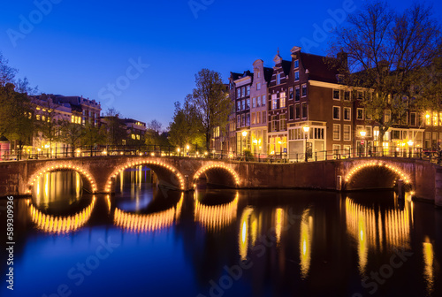 A tranquil evening in Amsterdam, illuminated by the soft glow of dusk. A beautiful arch bridge reflects on a canal amidst stunning architecture and river views. © Taljat