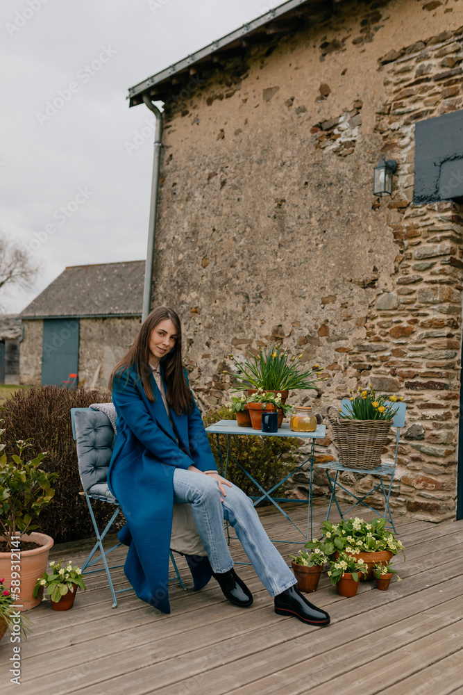 Full-length photo of pretty Caucasian girl with loose hair wearing blue coat, jeans and boots sitting on summer terrace with flowers and resting in spring weekend day.