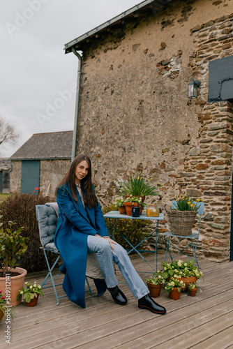 Obraz na płótnie Full-length photo of pretty Caucasian girl with loose hair wearing blue coat, jeans and boots sitting on summer terrace with flowers and resting in spring weekend day. w salonie