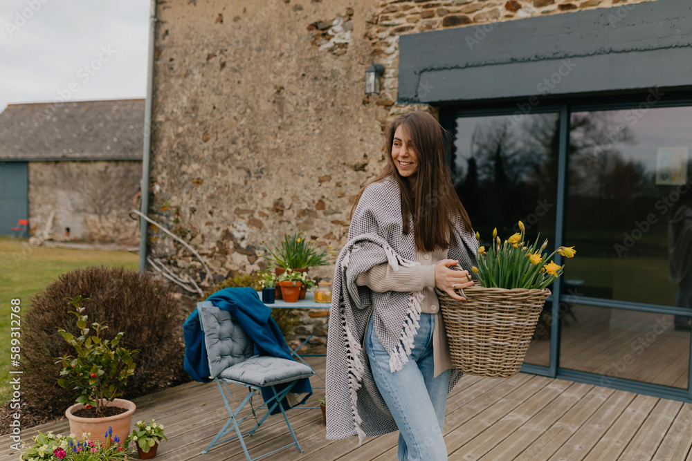 Fototapeta premium Smiling happy young woman with loose hair wearing jeans and throws a blanket on shoulders is standing on wooden terrace with big basket with flowers on background of green filed.