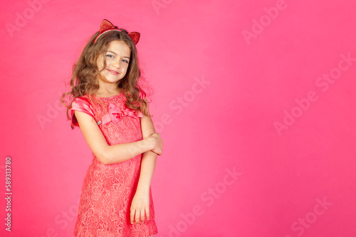 Portrait of happy smiling little girl in pink dress posing emotion at pink background, looking at camera. Funny blonde young lady expressing emotion. Childhood emotional concept. Copy ad text space
