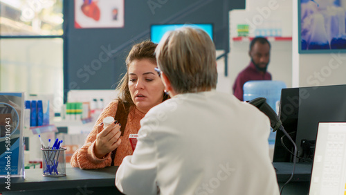 Young adult holding cardiology pills and talking to specialist at counter, asking about support to buy treatment. Buying boxes of vitamins and drugs at drugstore desk cash register.
