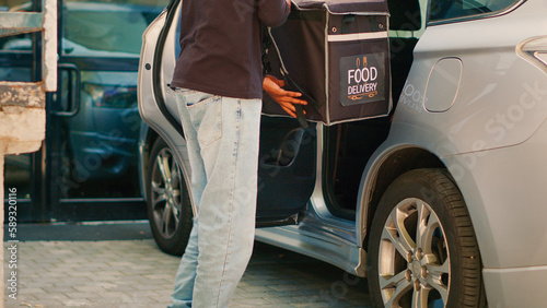 Restaurant employee taking backpack out of car to deliver fastfood meal, leaving vehicle to give food delivery order to client at front door. Takeaway courier delivering package. Handheld shot.