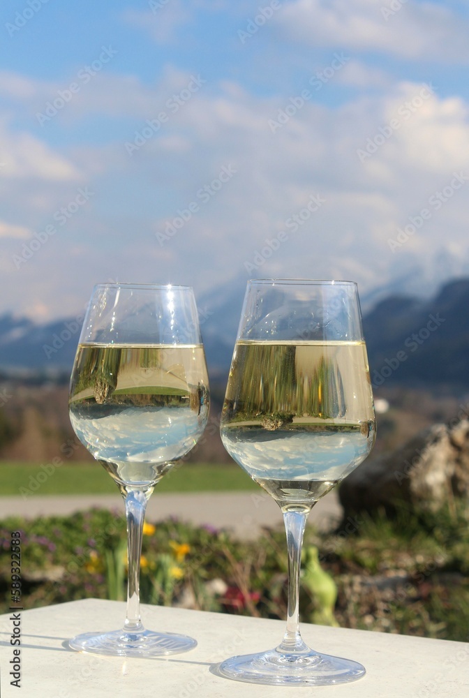 Zwei Gläser mit Weißwein. Im Hintergrund die Alpen. In den Gläsern spiegelt sich die Natur. Allgäu, Bayern, im Frühling Zwei Gläser mit Weißwein. Im Hintergrund die Alpen. In den Gläsern spiegelt sich die Natur. Allgäu, Bayern, im Frühling